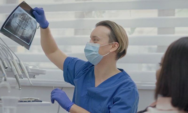 Dentist in blue scrubs and mask reviewing dental X-ray with a patient present